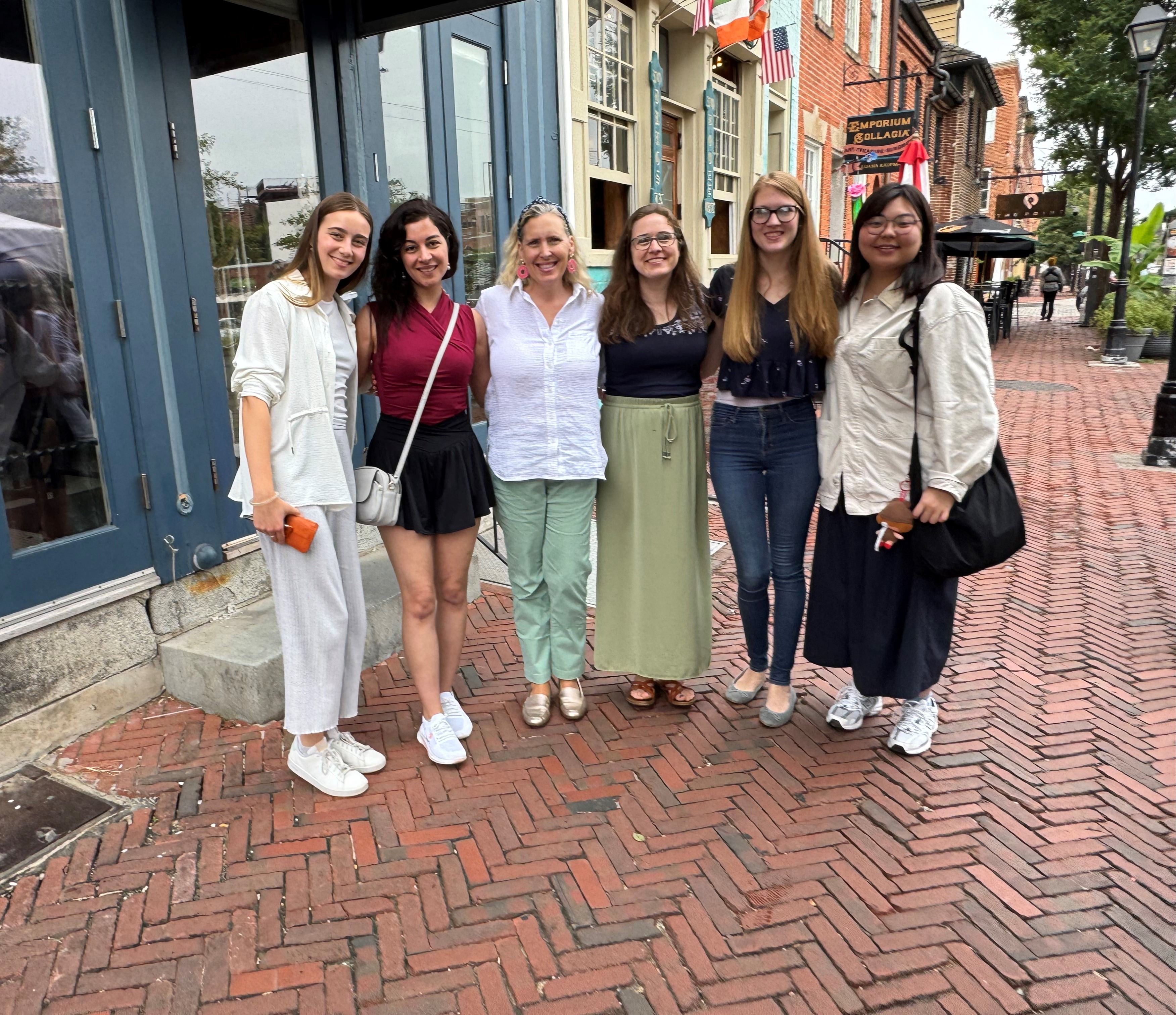 Main Image Description: Several women in various casual attire pose for a photo on a herringbone-patterned brick sidewalk....